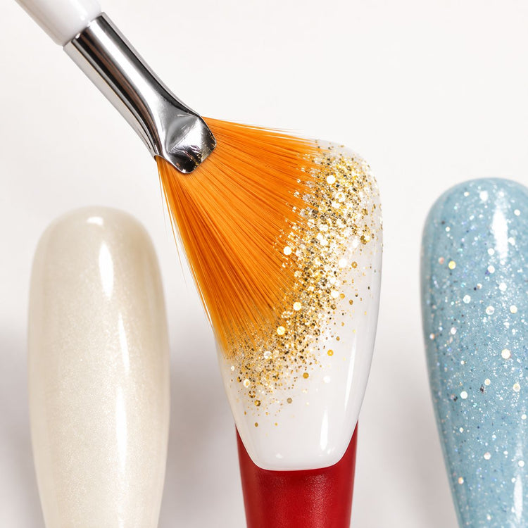 Orange fan brush sweeping gold glitter over a white gel nail tip, from the 20 piece nail art brush set