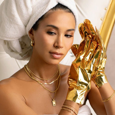 Lifestyle image of woman wearing VOESH Imperial Gold Collagen Gloves hand mask with towel-wrapped hair in a spa-style beauty setting