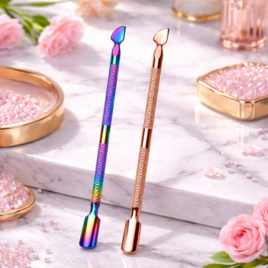 Rainbow and rose gold double-ended cuticle pushers and scrapers displayed on a marble beauty surface with pink roses and crystal accents