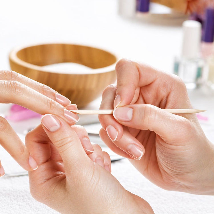 Close-up of a manicure in progress using a wooden cuticle pusher stick on natural nails, with nail care tools blurred in the background