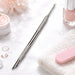 Stainless steel cuticle scraper and pusher with loop-detail tip and angled precision scraper, displayed on a manicure setup with nail polish, towel and nail file