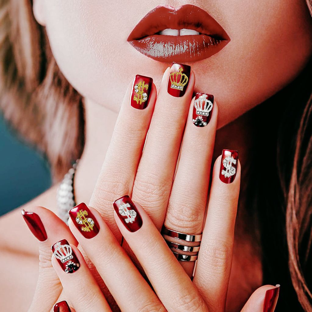 Burgundy gel nails with gold crown and dollar sign 3D nail charms and rhinestones—luxury manicure close-up
