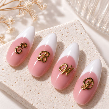 Four almond nails with gold script letter nail art charms on a textured stand