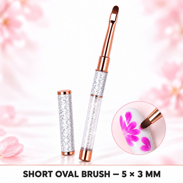 Short oval 5 × 3 mm nail art brush with crystal-filled handle and matching cap, shown painting pink flowers on a white nail.
