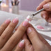 Nail tech patting blush pink gel onto an almond nail with a cube sponge held in a claw grabber tool