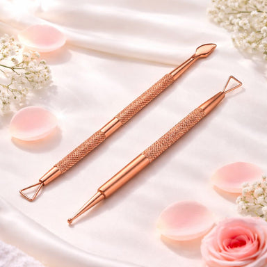 Rose gold double-ended cuticle scraper and loop tool with matching dotting and detail tool on a soft pink satin background with flower petals