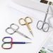 Lifestyle image of precision cuticle scissors in silver, gold and rainbow finishes arranged on a white textured surface beside stacked books